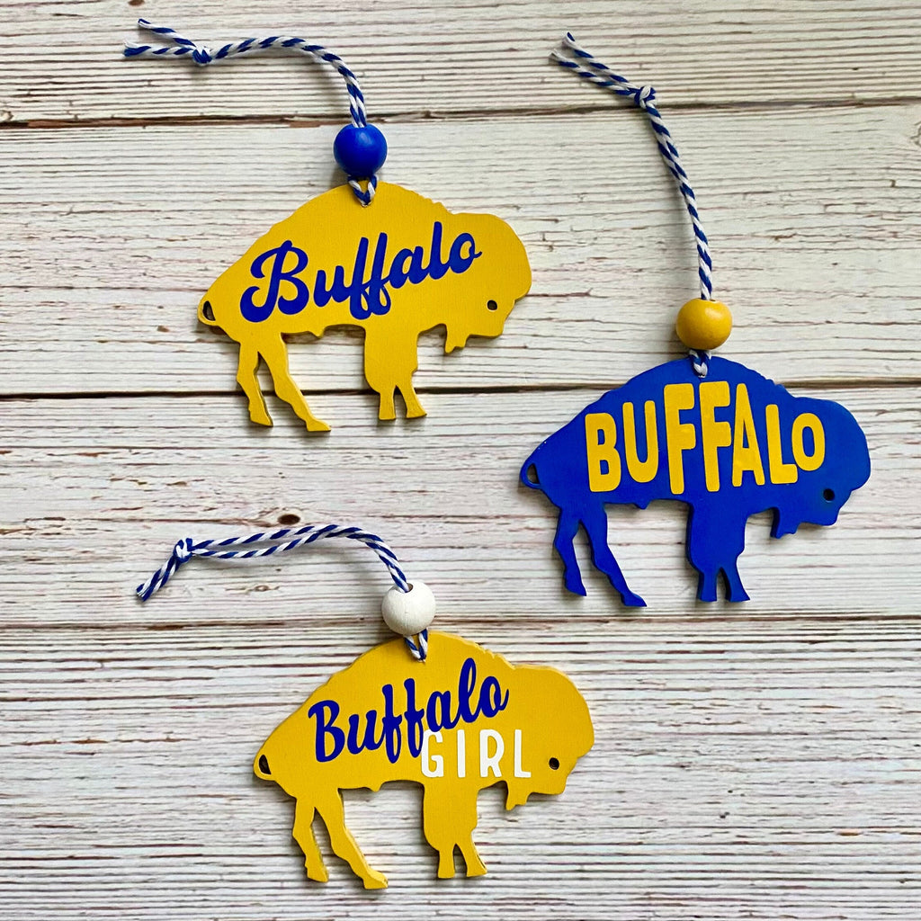 Three buffalo-shaped hanging ornaments on a whitewashed wood background. Two ornaments are yellow with blue 'Buffalo' lettering, and one is blue with yellow 'BUFFALO' lettering.