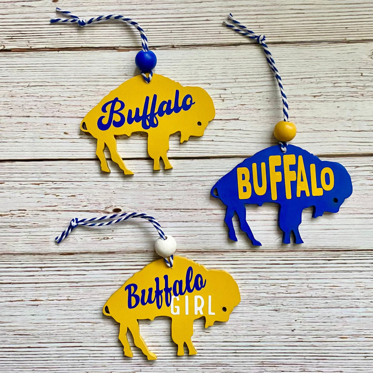 Three buffalo-shaped hanging ornaments on a whitewashed wood background. Two ornaments are yellow with blue 'Buffalo' lettering, and one is blue with yellow 'BUFFALO' lettering.