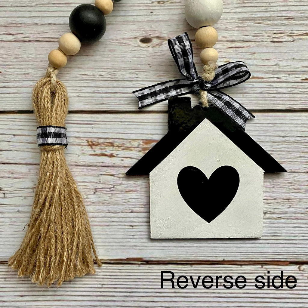 A decorative hanging ornament or wall hanging shown on a rustic whitewashed wood surface. The main visible item is the reverse side of a black-and-white beaded garland with a tassel, wooden beads, a black-and-white ribbon bow, and a house-shaped ornament with a black heart.