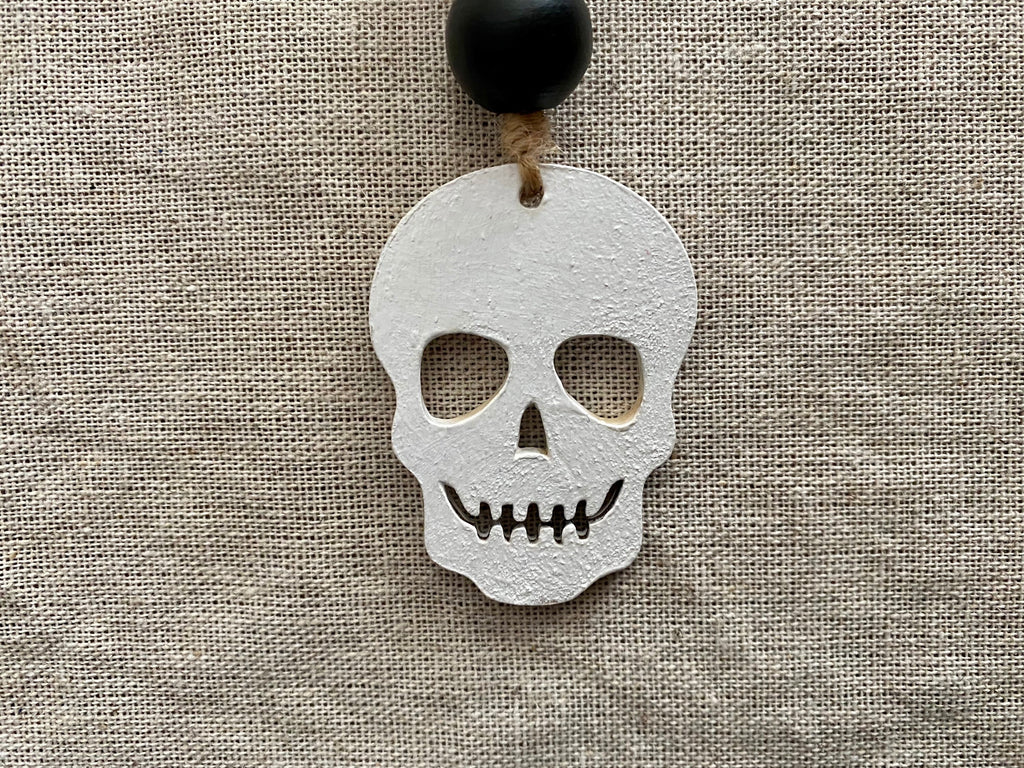 A white skull-shaped pendant with cutout eye sockets and a stitched mouth, hanging from a black bead on a beige linen background.