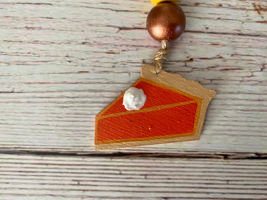 A close-up of a handcrafted wooden slice-of-pie charm with orange and red painted sections, a white cream detail, and a copper-colored bead above it on a whitewashed wood background.