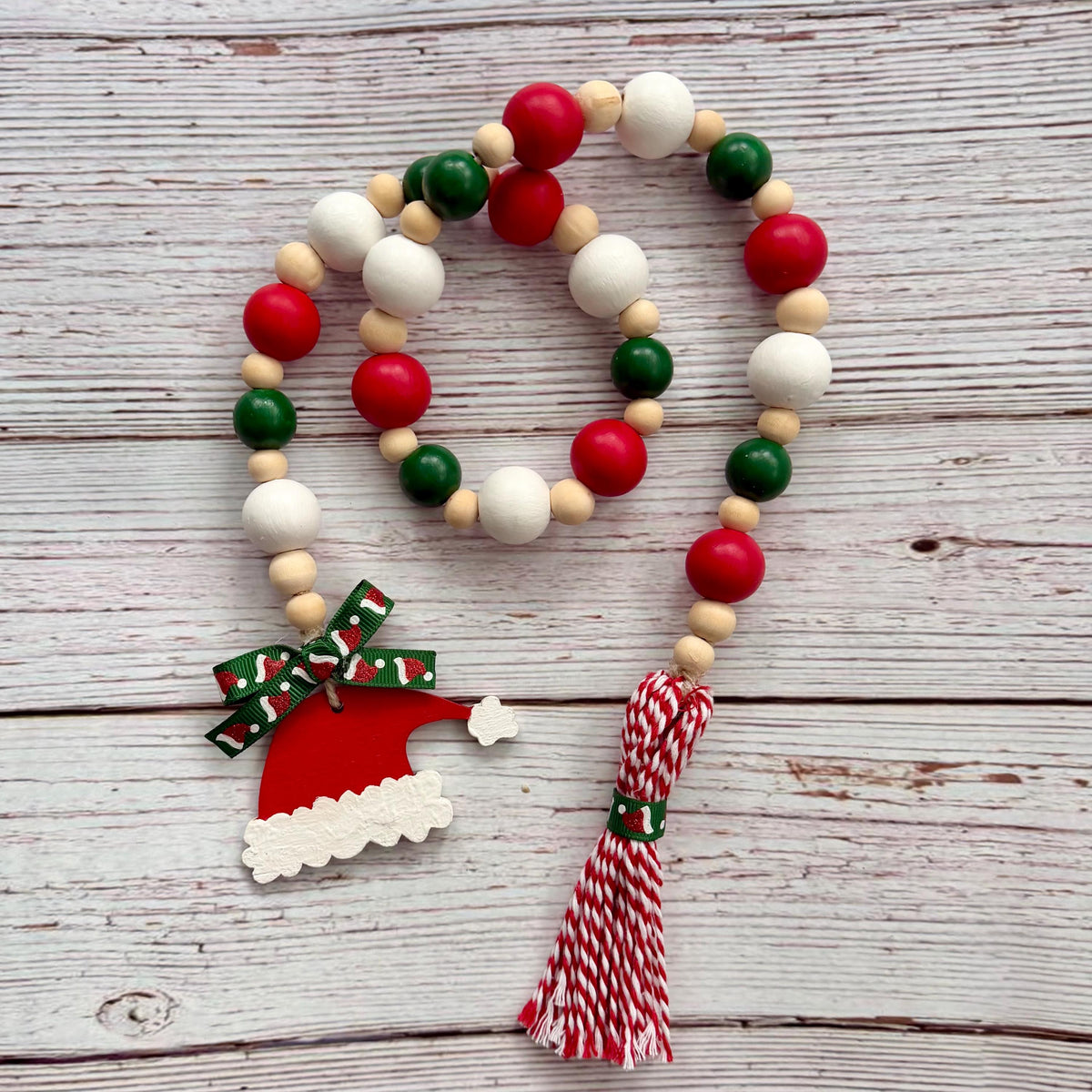 A Christmas-themed wooden bead garland with red, green, and white beads, a Santa hat ornament, a red and white patterned bow, and a red-and-white striped tassel, displayed on a rustic whitewashed wood background.