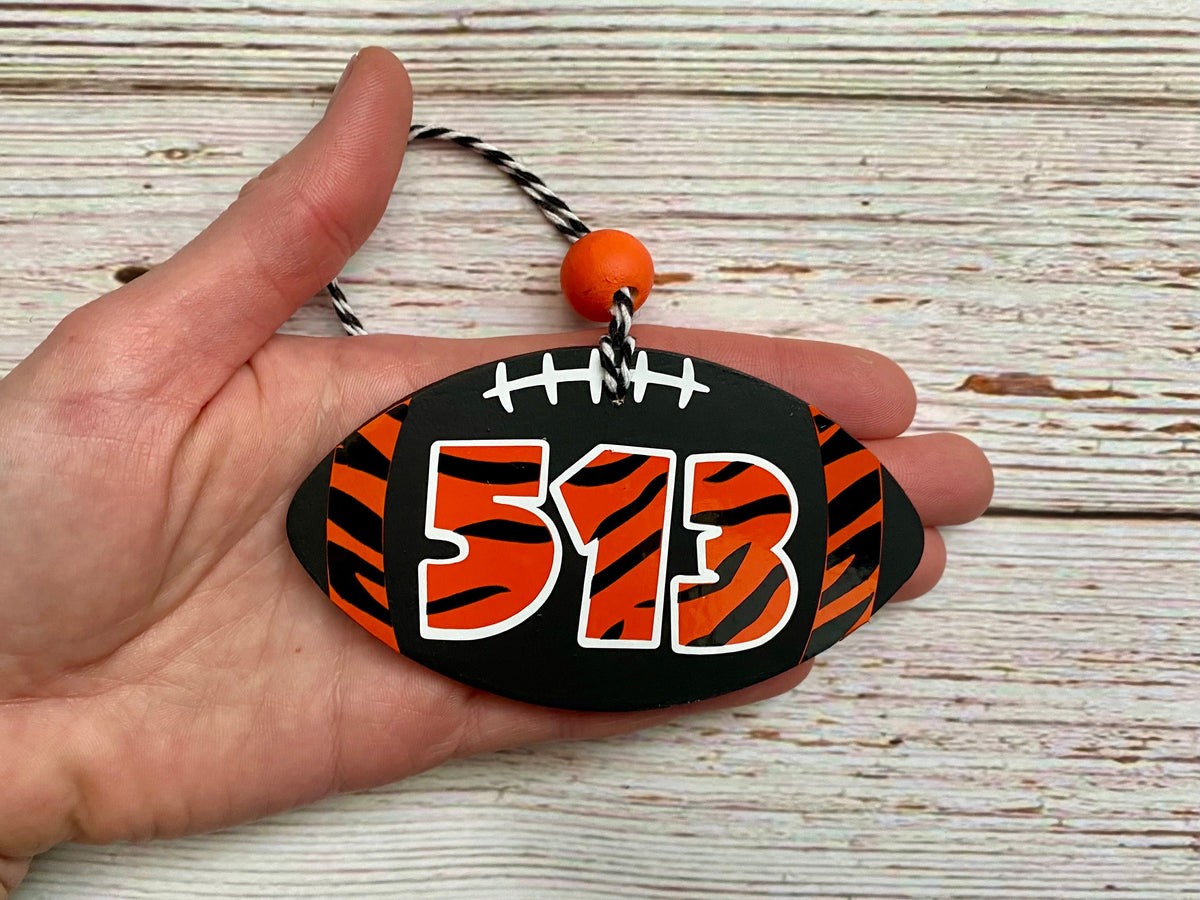 A hand holding a custom football-shaped ornament or plaque with the number 513 in orange and black tiger stripes, featuring a black and white striped hanging cord and an orange bead, set against a rustic white-washed wood background.