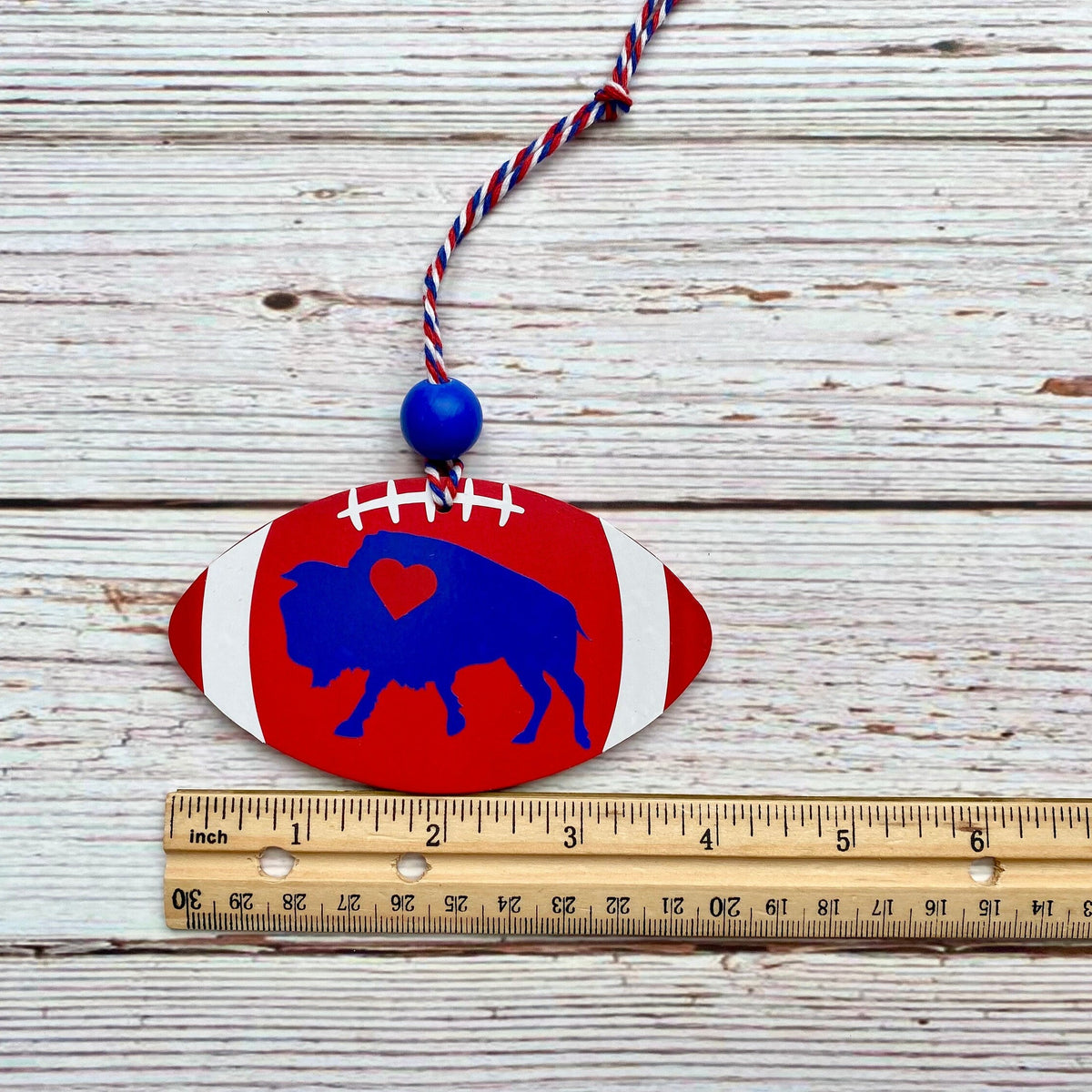 A hand-painted wooden football ornament with a blue buffalo silhouette, red and white stripes, and a red, white, and blue hanging cord, shown next to a wooden ruler on a whitewashed wood background.