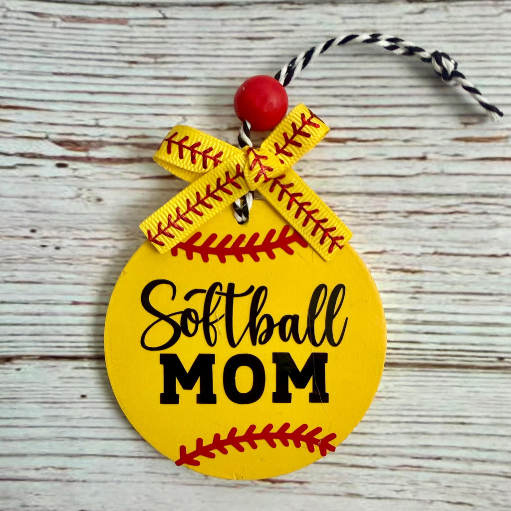 A round yellow softball-shaped ornament with 'Softball MOM' in black lettering, red seam details, a yellow ribbon bow, and a red bead on a rustic white wood background.