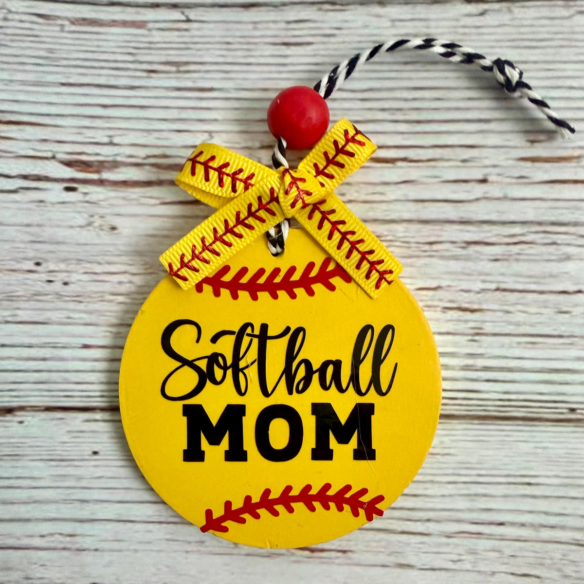 A round yellow softball-shaped ornament with 'Softball MOM' in black lettering, red seam details, a yellow ribbon bow, and a red bead on a rustic white wood background.