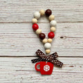 A beaded garland with natural wood beads, red and dark brown beads, and a brown polka dot ribbon bow tied around the bottom, featuring a red mug charm with a white snowflake, displayed on a rustic white-washed wooden background.