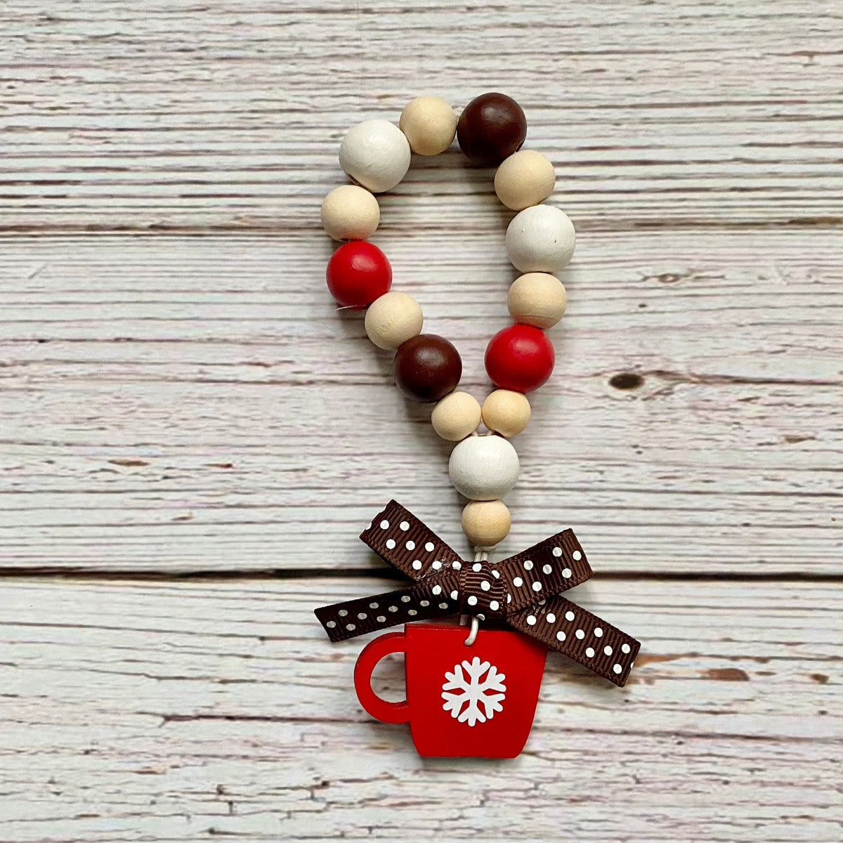 A beaded garland with natural wood beads, red and dark brown beads, and a brown polka dot ribbon bow tied around the bottom, featuring a red mug charm with a white snowflake, displayed on a rustic white-washed wooden background.