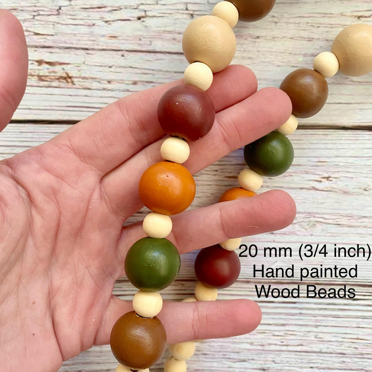 A hand holds a strand of hand-painted wooden beads in natural beige, brown, orange, and green colors over a rustic white wood plank background. Text on the image reads '20 mm (3/4 inch) Hand painted Wood Beads'.