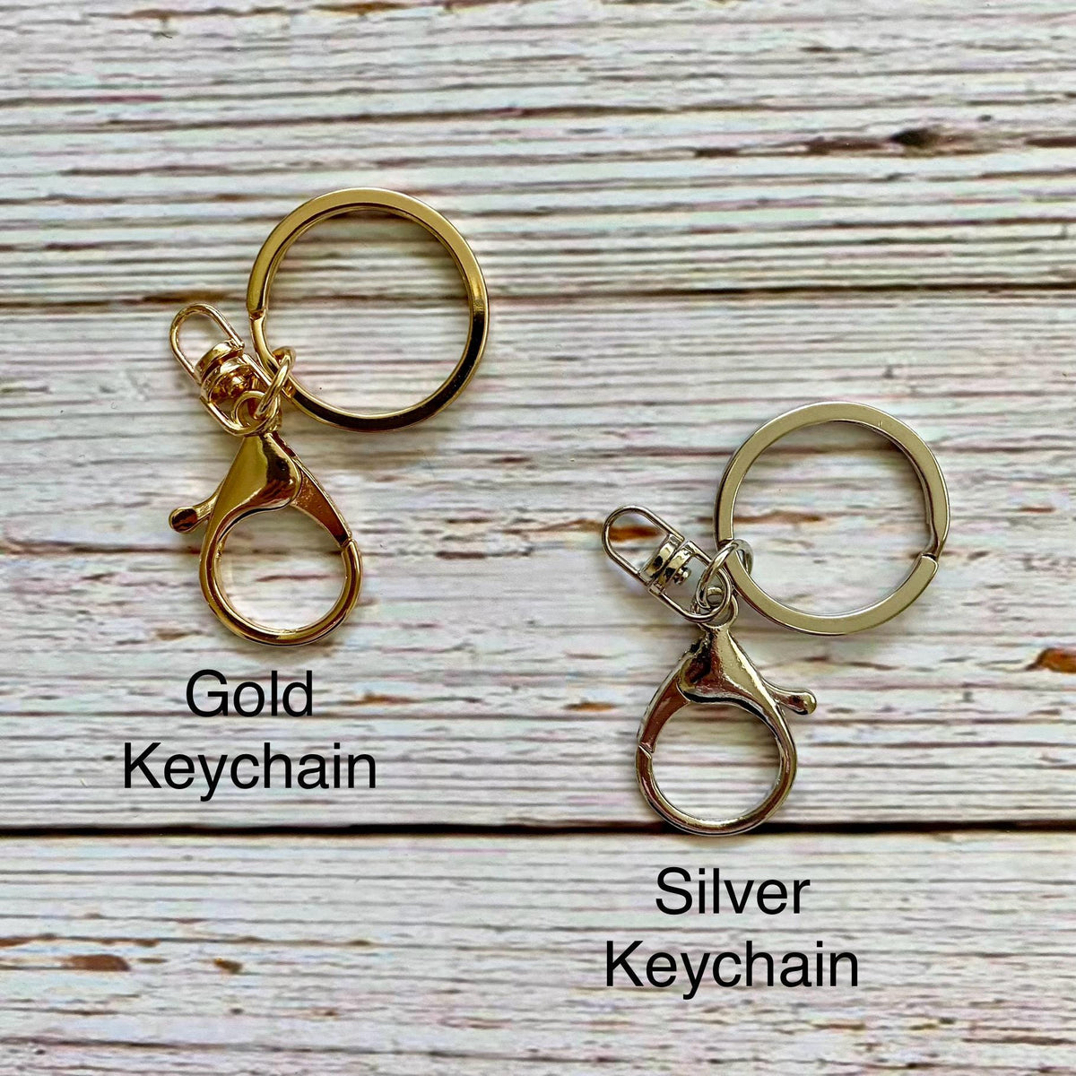 Two metal keychains shown on a whitewashed wood background, labeled 'Gold Keychain' and 'Silver Keychain.' The left keychain is gold-toned and the right keychain is silver-toned, both with split rings and lobster clasp attachments.