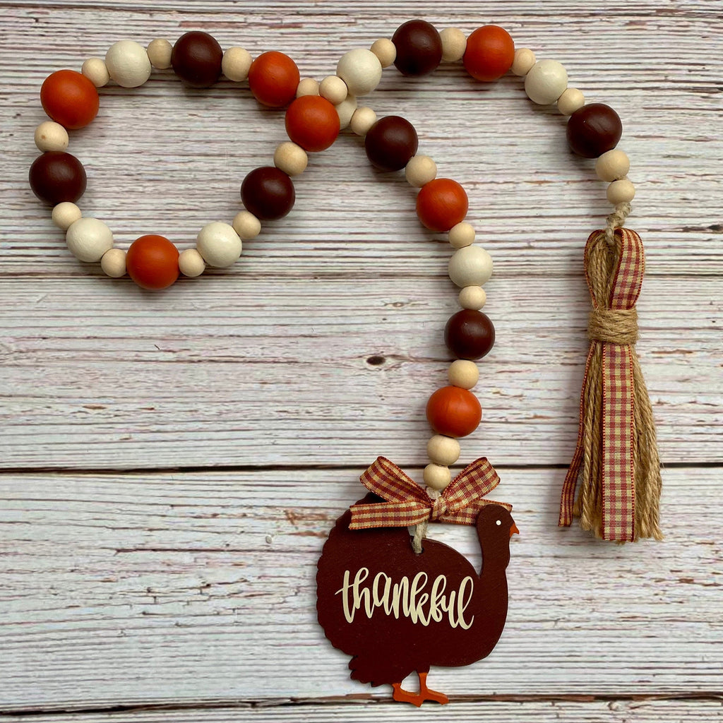 A fall-themed wooden bead garland with orange, dark brown, and cream beads, featuring a tassel and a turkey-shaped wooden ornament that reads 'thankful' in white script, displayed on a rustic white wood plank background.