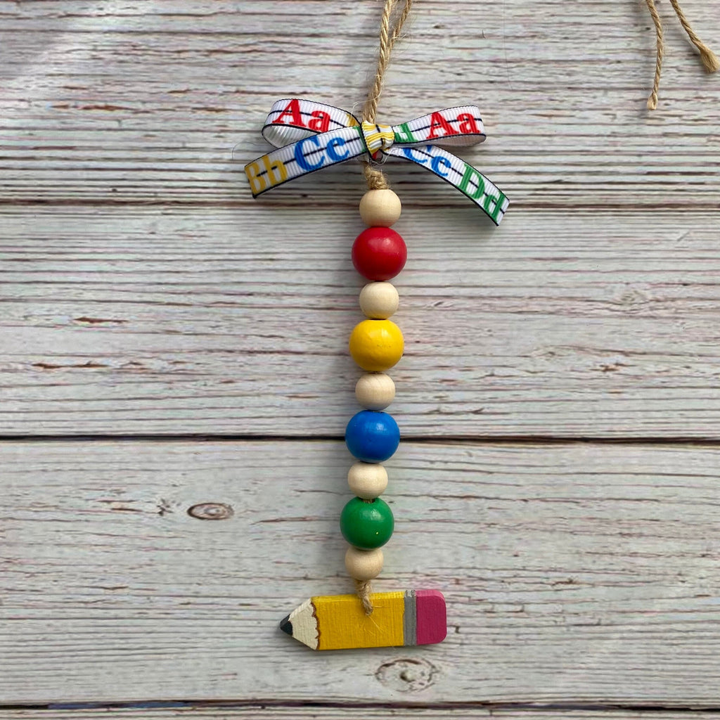 A handmade educational bead garland ornament featuring colorful alphabet beads, wooden beads, a red bead, a yellow bead, a blue bead, a green bead, and a small pencil charm at the bottom, tied with a patterned ribbon on a rustic wood background.