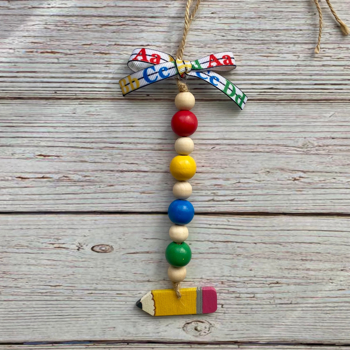 A handmade educational bead garland ornament featuring colorful alphabet beads, wooden beads, a red bead, a yellow bead, a blue bead, a green bead, and a small pencil charm at the bottom, tied with a patterned ribbon on a rustic wood background.