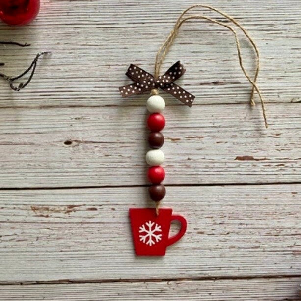 A rustic Christmas ornament featuring a red mug with a white snowflake, decorated with red, white, and brown wooden beads, a brown polka-dot bow, and a jute hanging string, displayed on a weathered light gray wood plank background.
