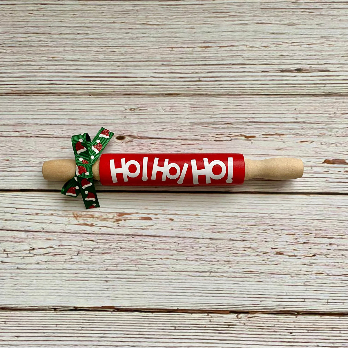 A wooden rolling pin with a red painted center wrapped in white 'Ho Ho Ho' lettering and tied with a green holiday ribbon, placed on a whitewashed wooden plank surface.