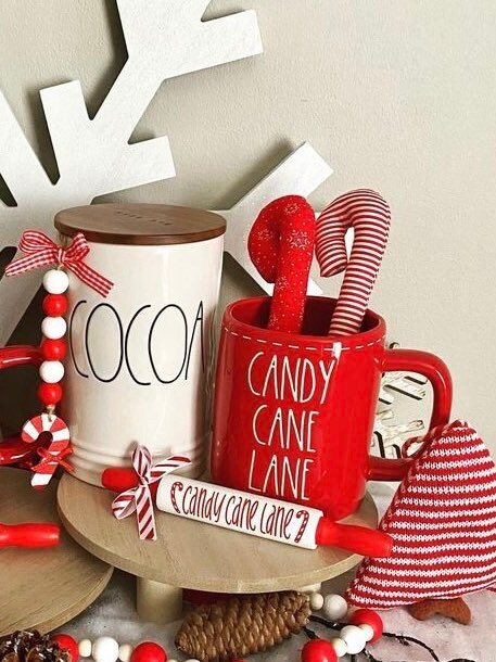 A styled holiday tabletop scene with two mugs and canisters: a white ceramic canister labeled COCOA with a wooden lid and red-and-white bead garland, and a red ceramic mug labeled CANDY CANE LANE with red candy canes inside. A small sign reads candy cane lane, with red-and-white striped fabric and holiday decor around it on a light neutral background.