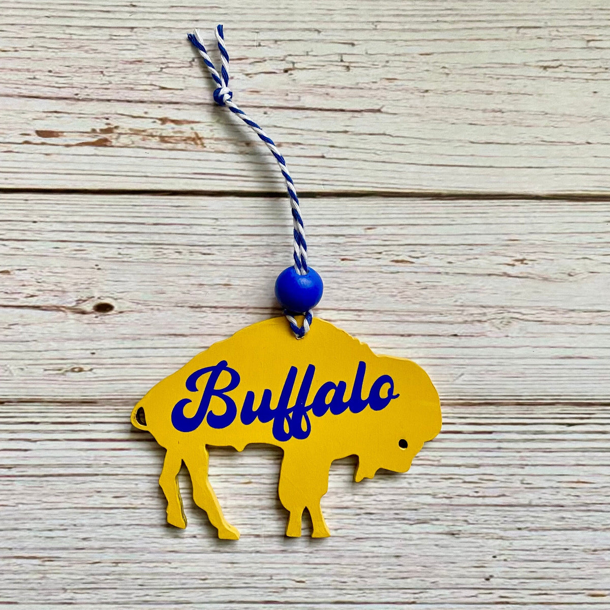 A yellow bison-shaped ornament with the word 'Buffalo' in blue script, hanging from a blue-and-white string on a whitewashed wood plank background.
