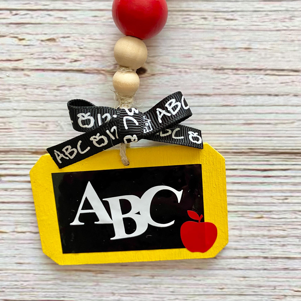 A personalized school-themed ornament featuring a yellow chalkboard-shaped tag with 'ABC' in white letters on a black background, a small red apple, and a black ribbon printed with white letters. The ornament is decorated with red, gold, and natural wood beads at the top, set against a light-colored wooden background.
