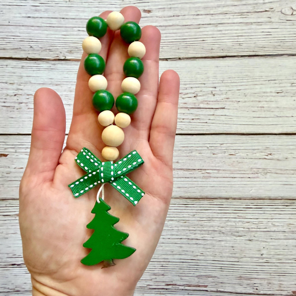 A hand holding a beaded Christmas ornament strand with green and cream wooden beads, a green ribbon bow, and a green Christmas tree charm, set against a rustic white wood plank background.