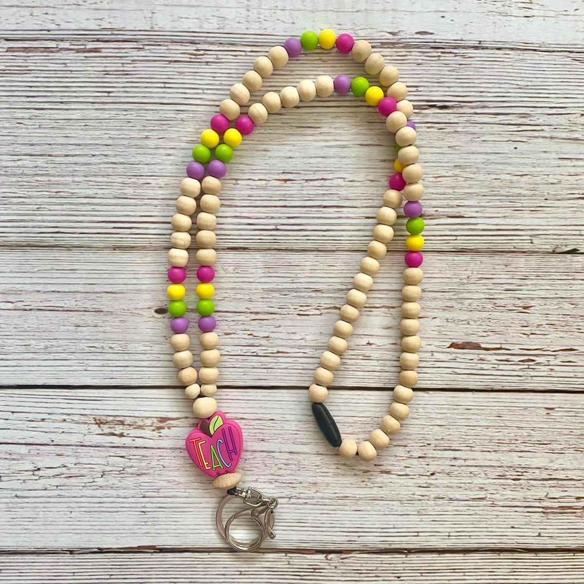 A beaded keychain or lanyard made of natural wood beads with colorful accents in pink, green, yellow, and purple, featuring a pink heart charm with colorful stripes and a silver metal clasp, displayed on a rustic white-washed wooden background.