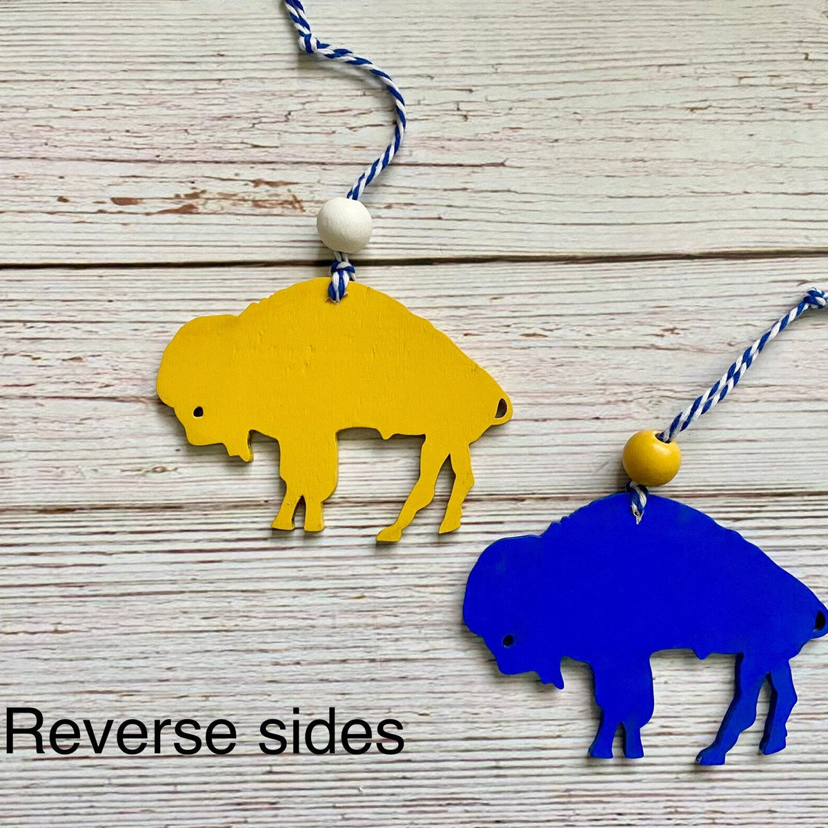 Two buffalo-shaped hanging ornaments in yellow and blue, each with a wooden bead and blue-and-white string, displayed on a whitewashed wood background. Text in the lower left reads 'Reverse sides'.