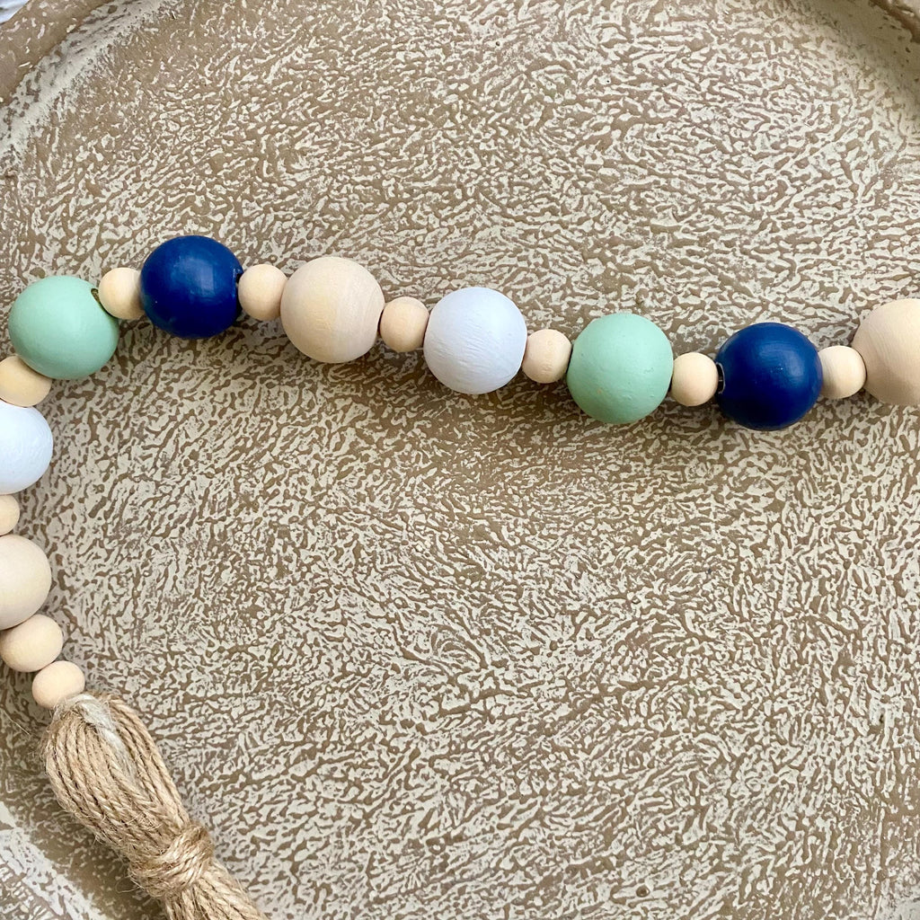 A wooden bead garland with large and small beads in navy blue, mint green, white, and natural wood, finished with a knotted tassel, displayed on a textured beige tray.