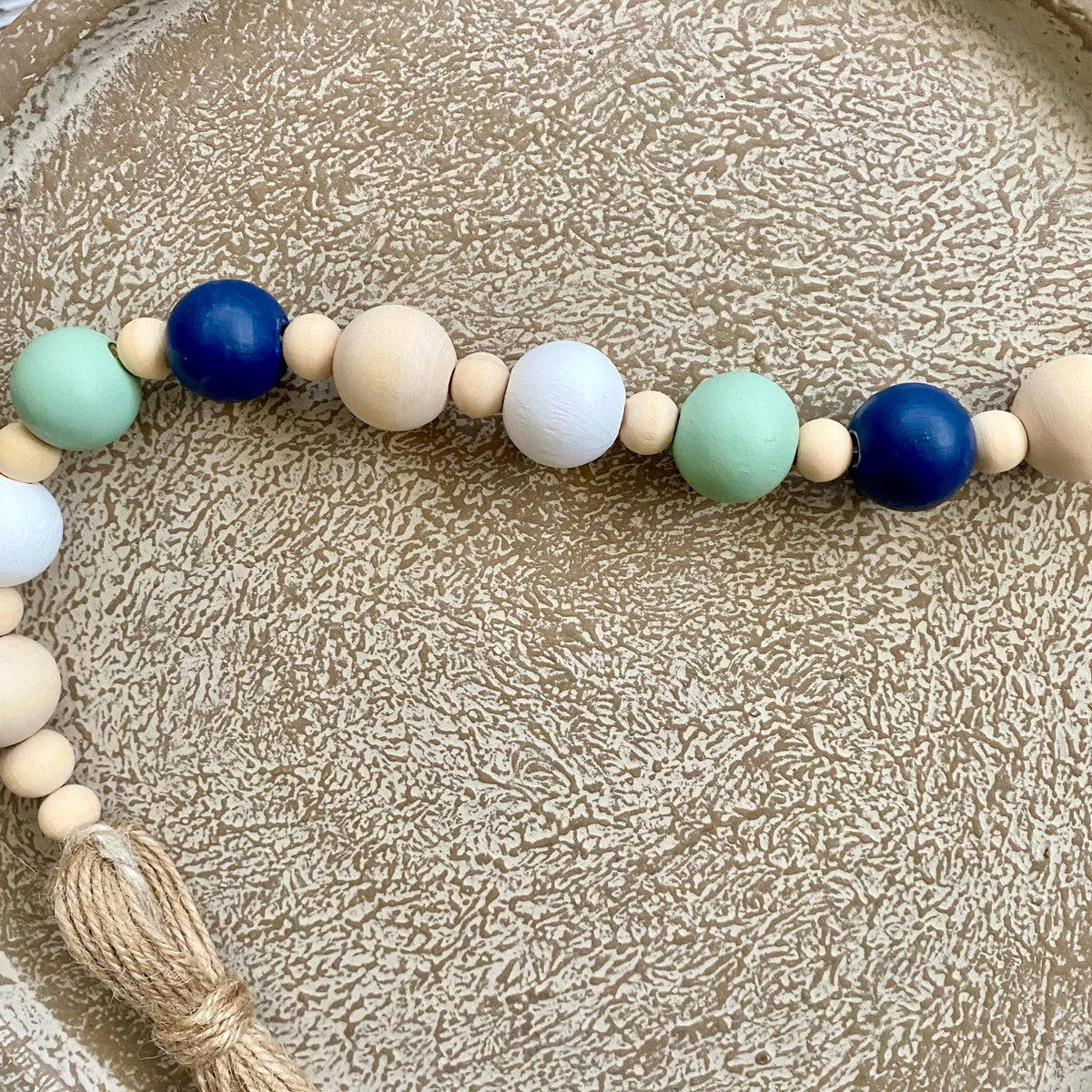 A wooden bead garland with large and small beads in navy blue, mint green, white, and natural wood, finished with a knotted tassel, displayed on a textured beige tray.