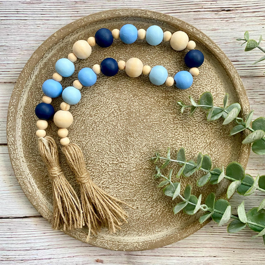 A beaded tassel garland with light blue, navy, and natural wood beads, displayed on a round textured gold tray with eucalyptus leaves.