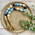 A beaded tassel garland with light blue, navy, and natural wood beads, displayed on a round textured gold tray with eucalyptus leaves.