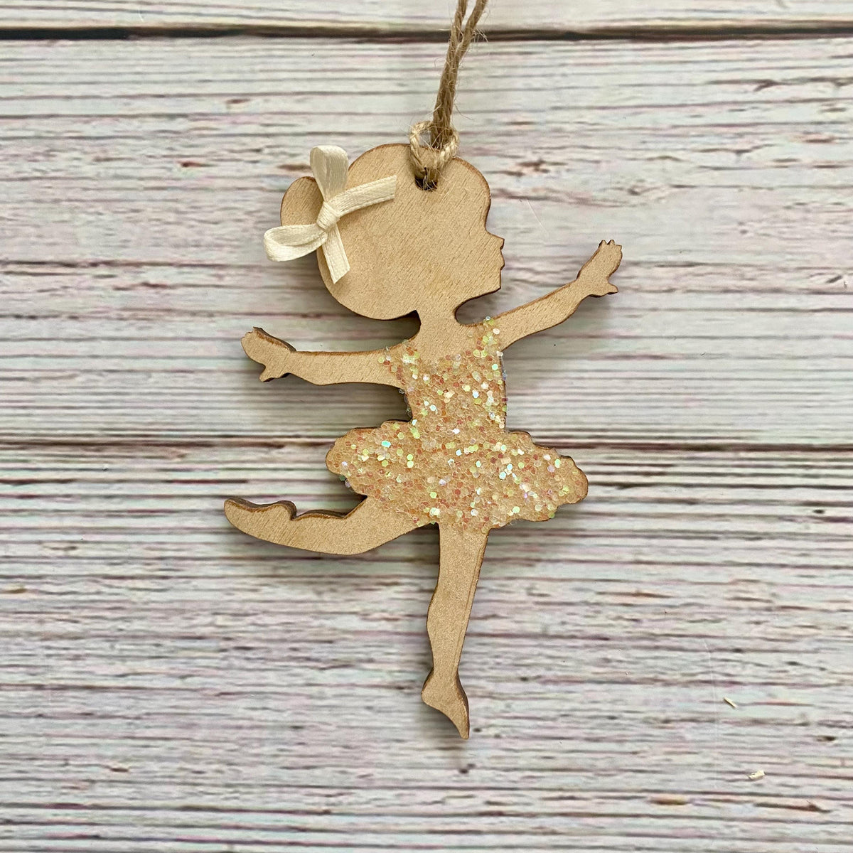 A wooden ballerina silhouette ornament with a glittery gold dress and a small cream ribbon bow, hanging on a rustic whitewashed wood background.