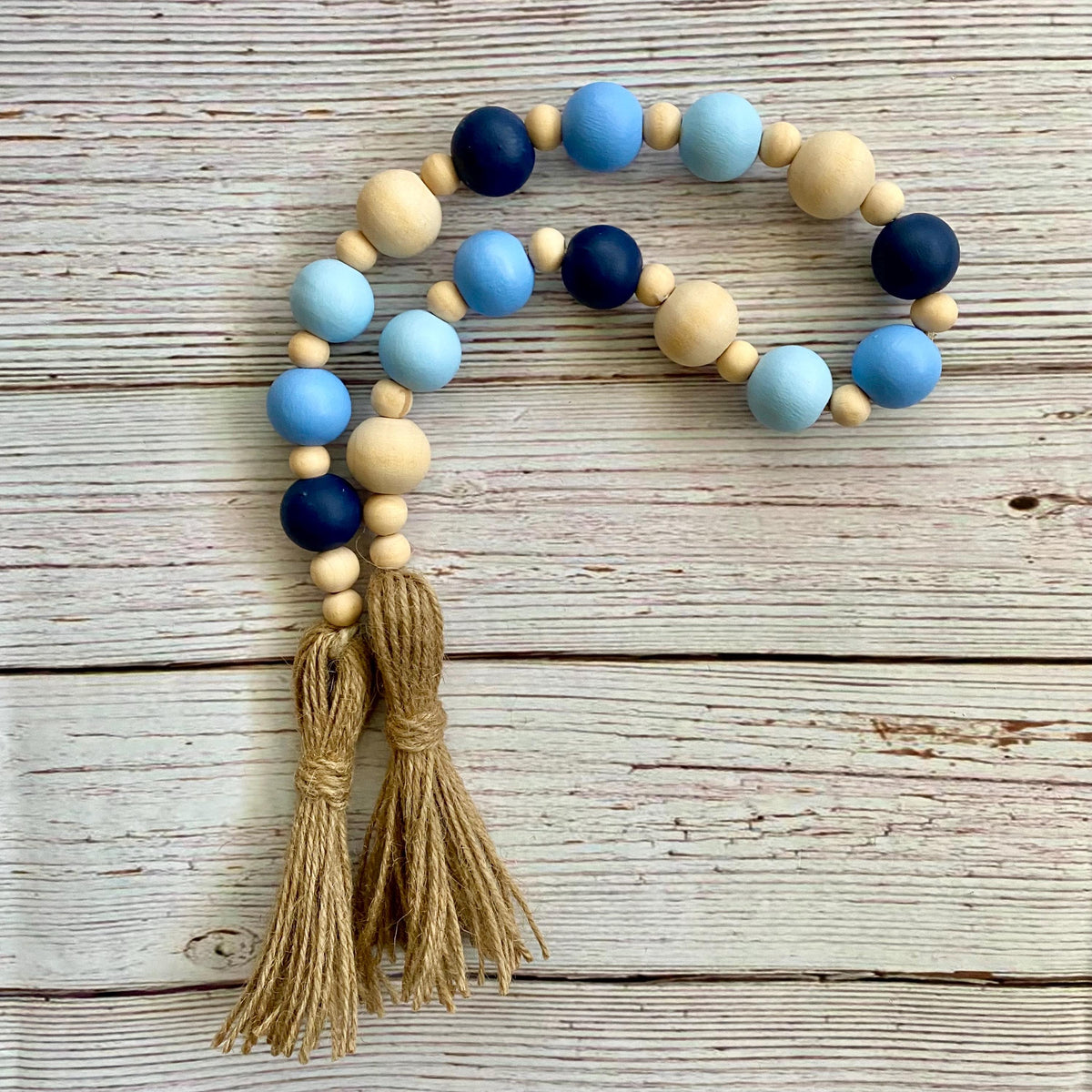 A beaded garland with light blue, navy, and natural wood beads, featuring two tan jute tassels, displayed on a rustic whitewashed wooden background.