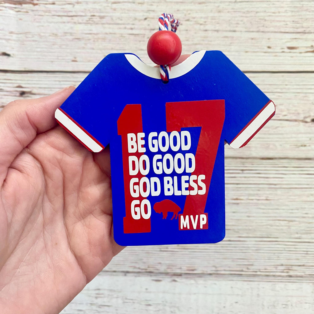 A hand holds a blue wooden ornament shaped like a sports jersey with red and white trim. The ornament features the number 17 in red and white block letters, with the text 'BE GOOD DO GOOD GOD BLESS GO MVP' in white.