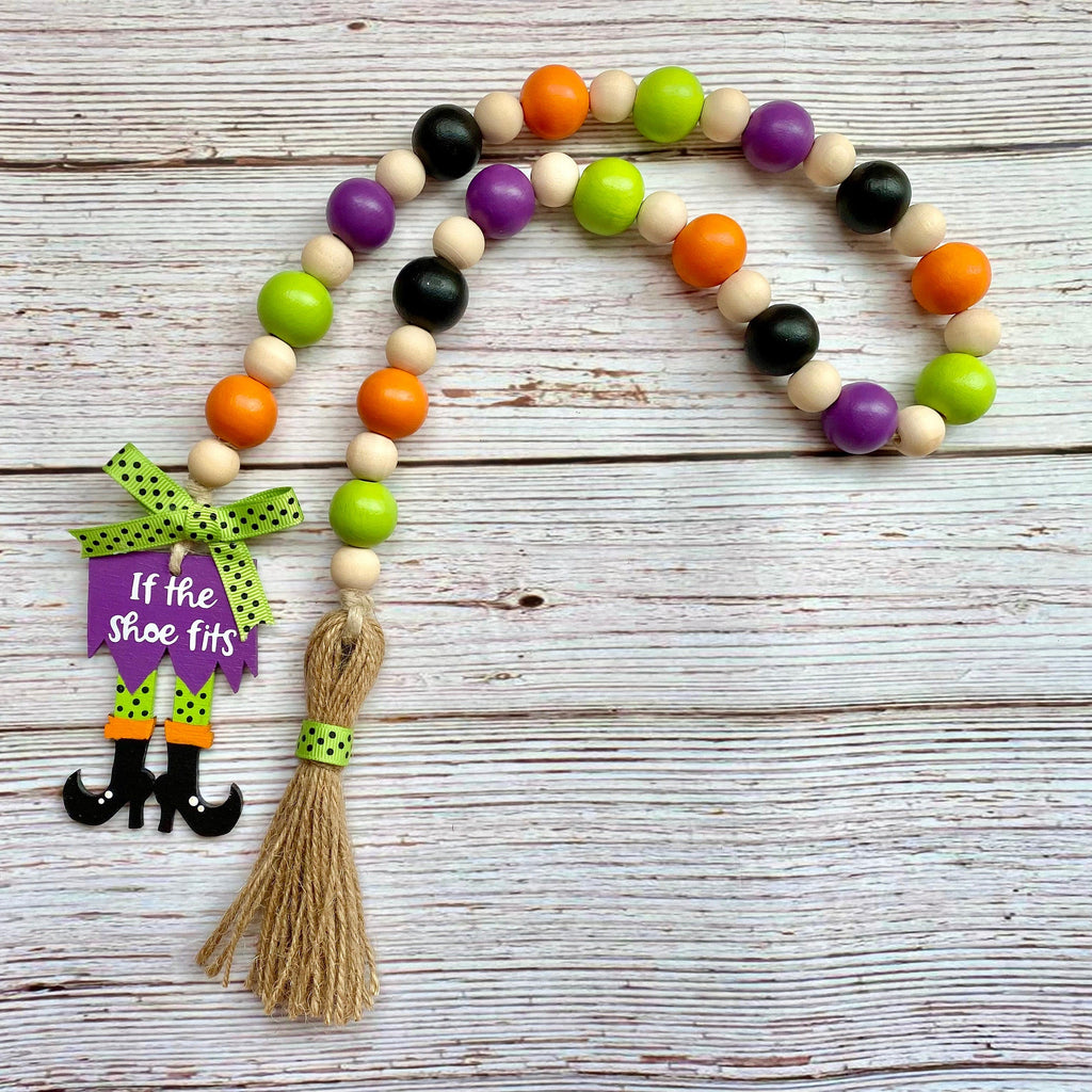 A Halloween-themed wooden bead garland with orange, green, purple, and black beads, a purple 'If the shoe fits' sign, and a jute tassel, displayed on a white-washed wooden background.