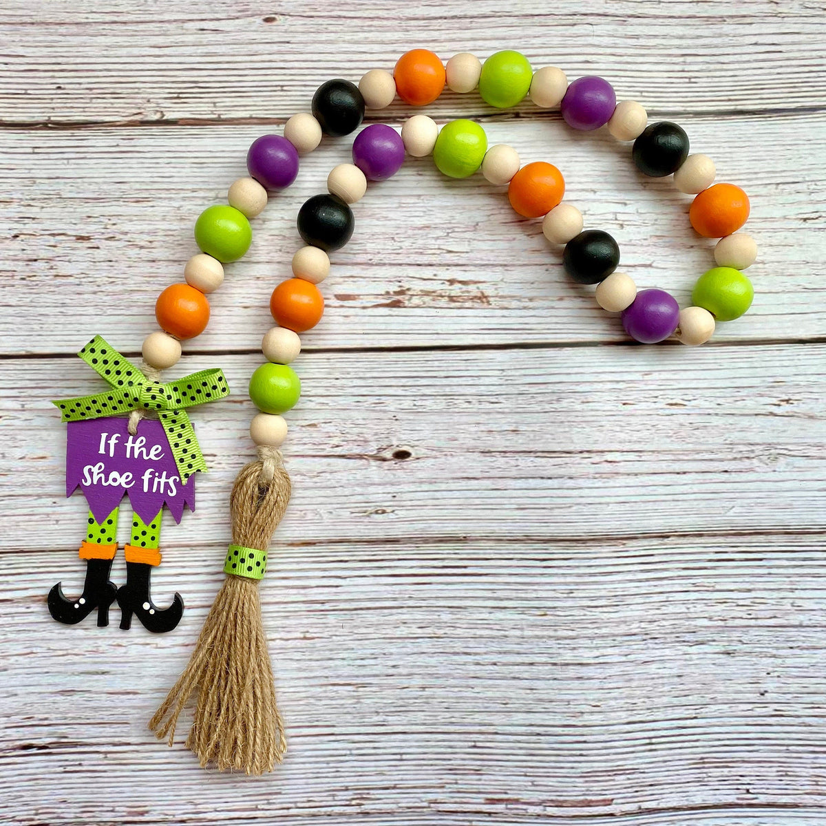 A Halloween-themed wooden bead garland with orange, green, purple, and black beads, a purple 'If the shoe fits' sign, and a jute tassel, displayed on a white-washed wooden background.