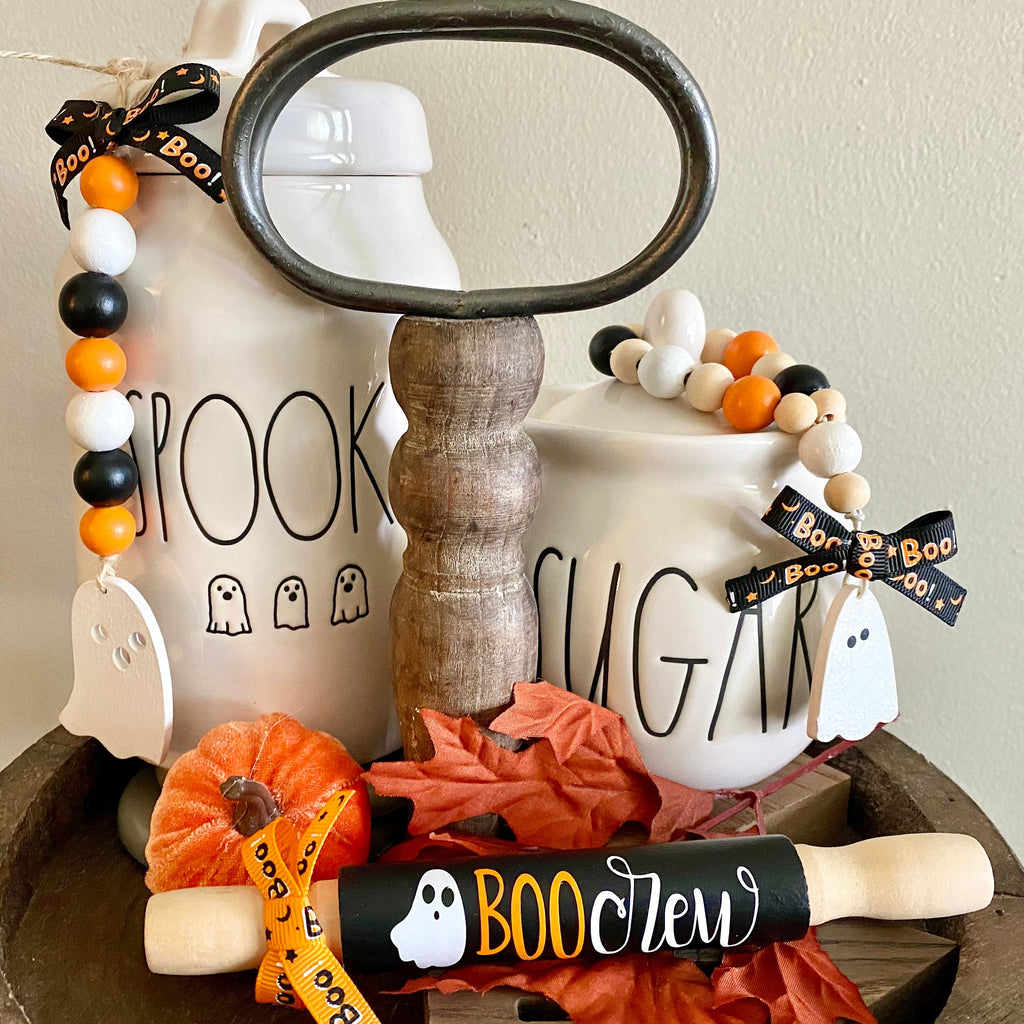 A styled Halloween-themed decorative arrangement in a round wooden tray, featuring two white ceramic canisters labeled 'BROOKS' and 'SUGAR' with ghost cutouts, orange and black beads, a magnifying glass, a small pumpkin, autumn leaves, and a black-and-orange striped straw.