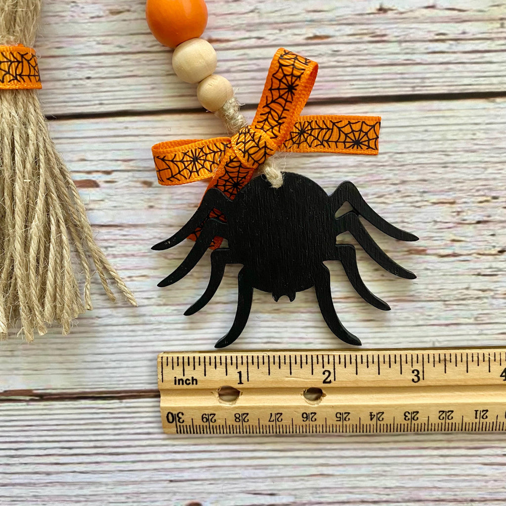 A black wooden spider ornament with an orange and black spiderweb ribbon bow, hanging next to a wooden ruler and a decorative broom on a whitewashed wood background.
