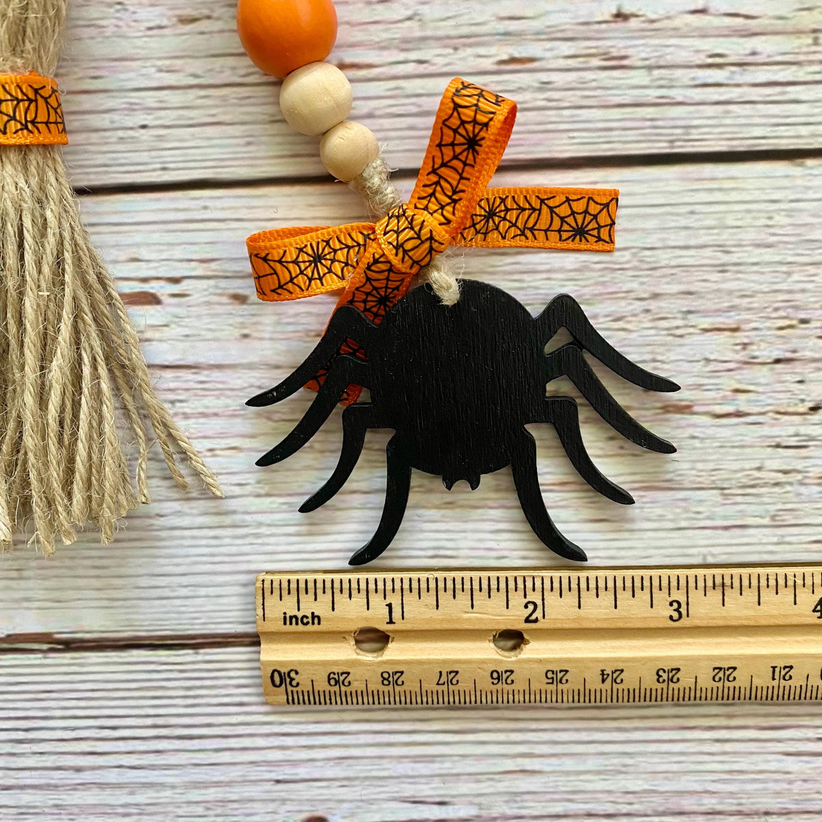A black wooden spider ornament with an orange and black spiderweb ribbon bow, hanging next to a wooden ruler and a decorative broom on a whitewashed wood background.