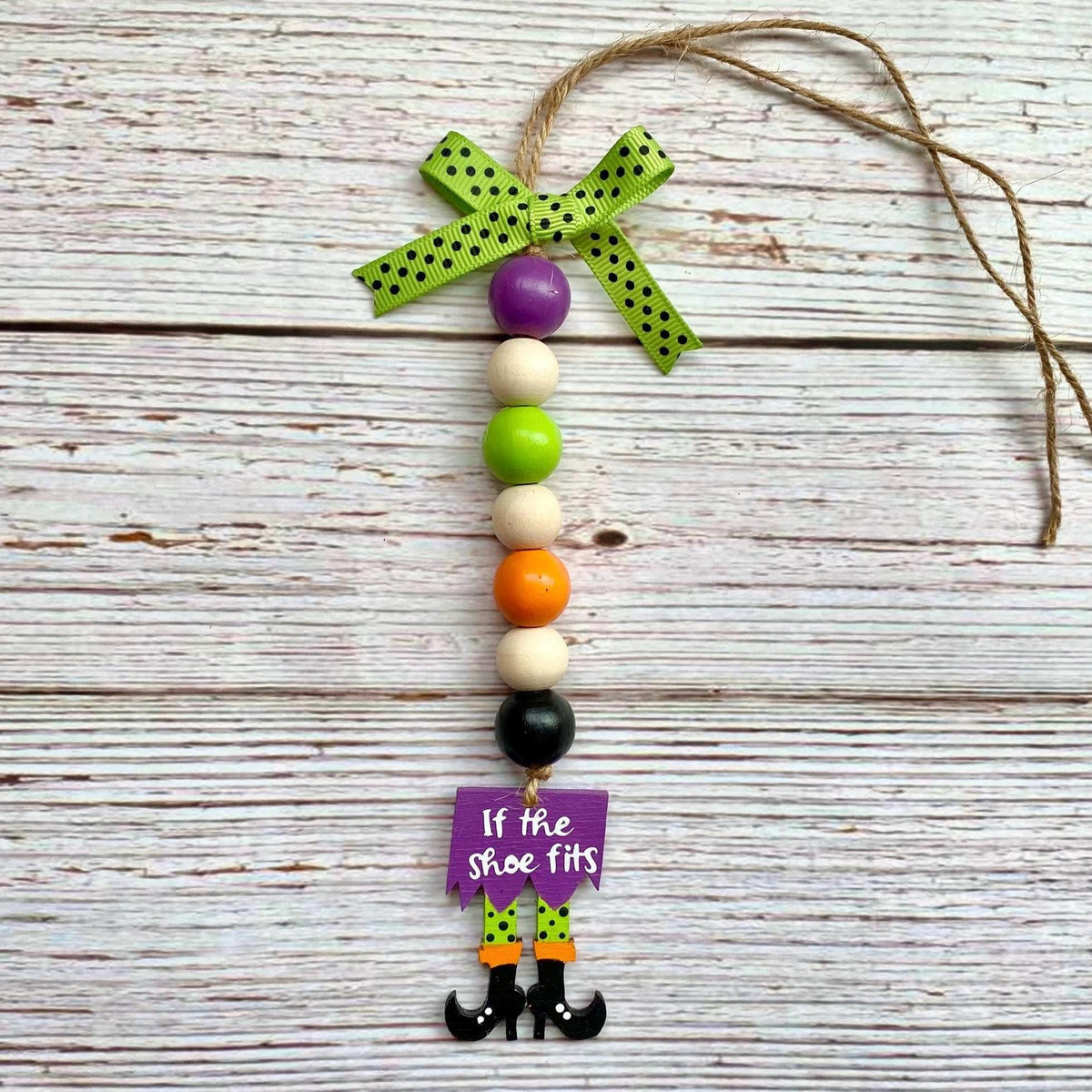 A Halloween hanging ornament made of wooden beads and a ribbon bow, with a purple tag that reads 'If the shoe fits' and small witch boot charms at the bottom, displayed on a whitewashed wood background.