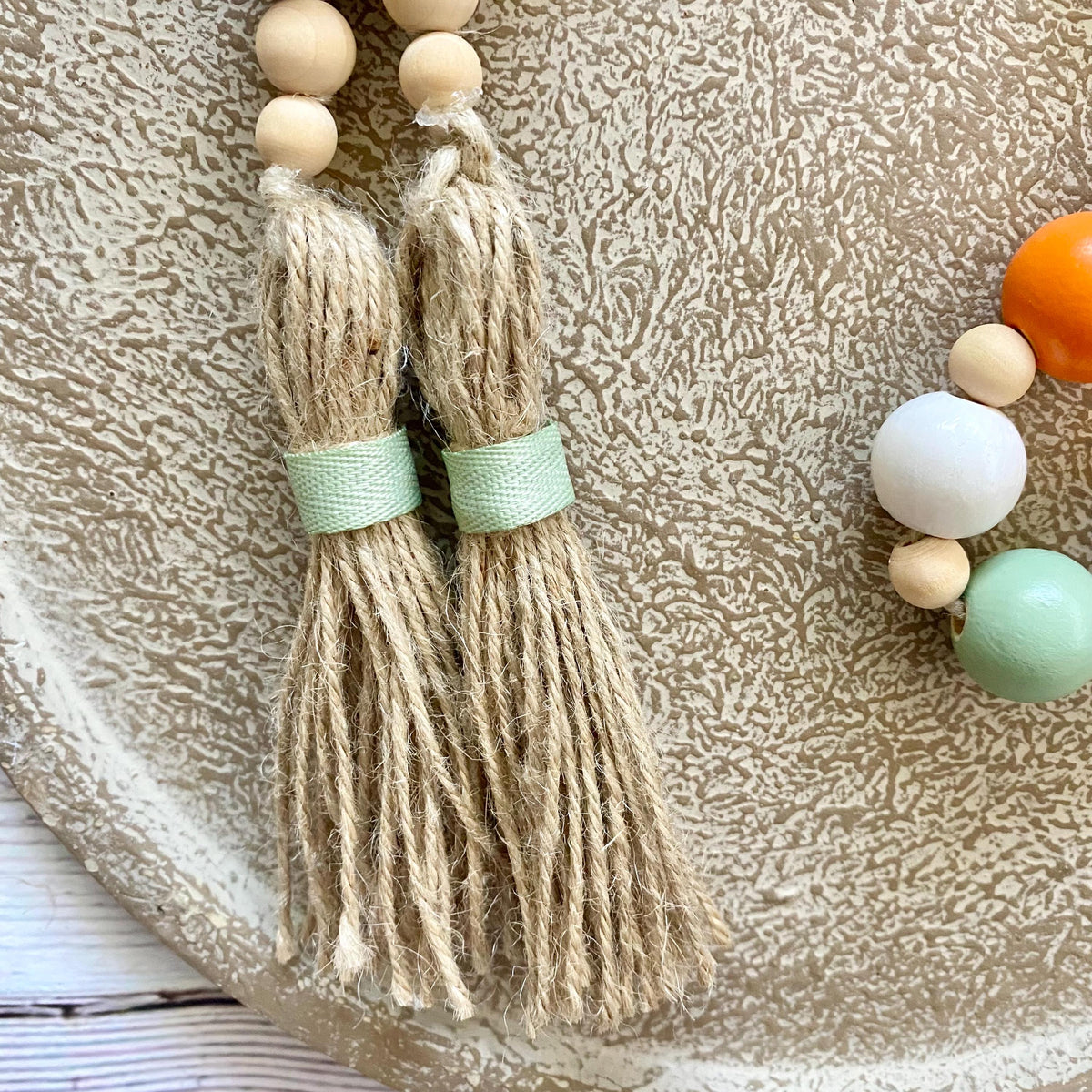Boho-style tassel garland with natural jute tassels, light wood beads, and mint green bands, displayed on a textured beige ceramic plate.