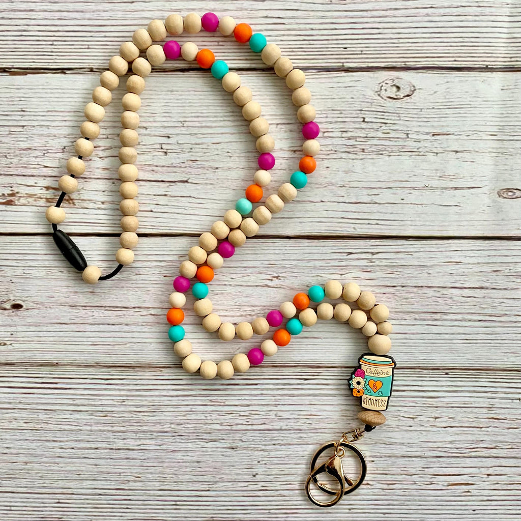 A wooden bead keychain necklace with colorful accent beads, a small 'caffeine & kindness' charm, and split keyrings, displayed on a rustic white-washed wood background.