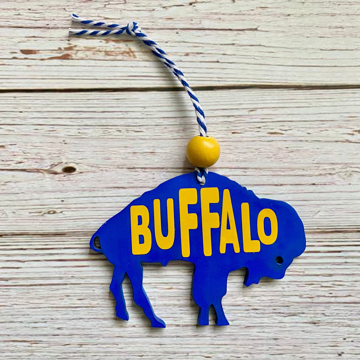 A blue buffalo-shaped hanging ornament with the word BUFFALO in yellow across the front, suspended by a blue-and-white string with a yellow bead, photographed on a whitewashed wood plank background.