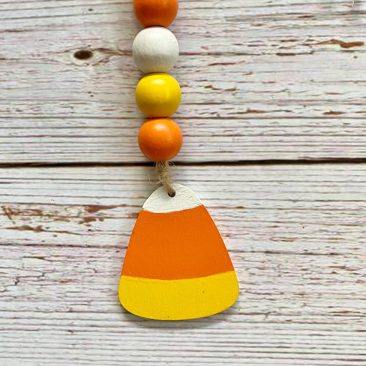 A wooden bead garland featuring orange, white, and yellow beads with a painted wooden candy corn charm, displayed on a rustic white-washed wood background.