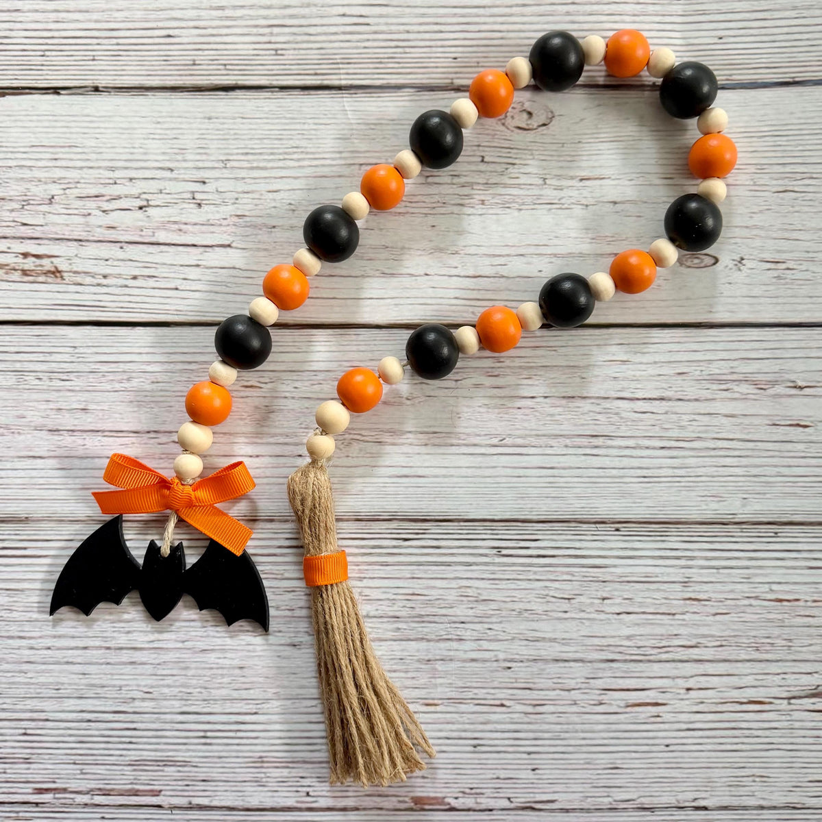 A Halloween-themed wooden bead garland with black and orange beads, a black bat cutout with an orange ribbon bow, and a beige jute tassel wrapped in orange ribbon, displayed on a white wood plank background.