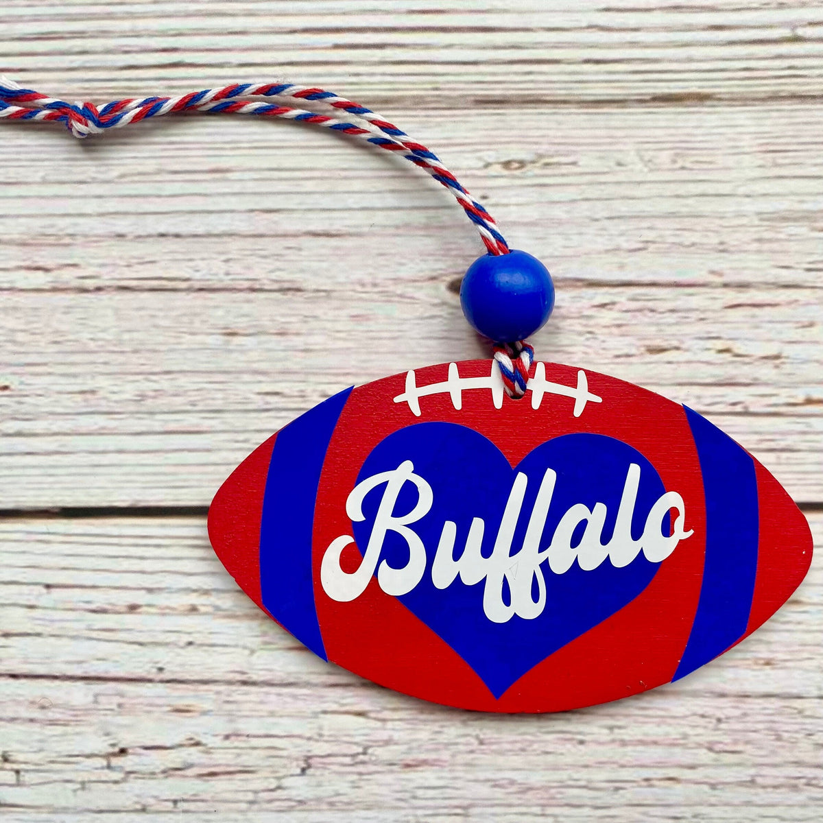 A custom wooden football ornament painted with red and blue stripes, a blue heart, and the word 'Buffalo' in white script, hanging from a red, white, and blue cord on a whitewashed wood background.