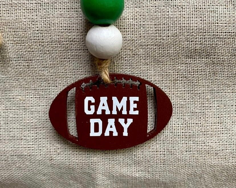 A wooden football-shaped ornament with 'GAME DAY' painted in white letters, hanging from a rope with a white bead, set against a beige burlap background.