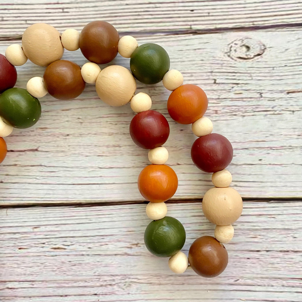 A wooden bead garland with large and small round beads in natural beige, brown, orange, red, and green, arranged on a whitewashed wooden surface.