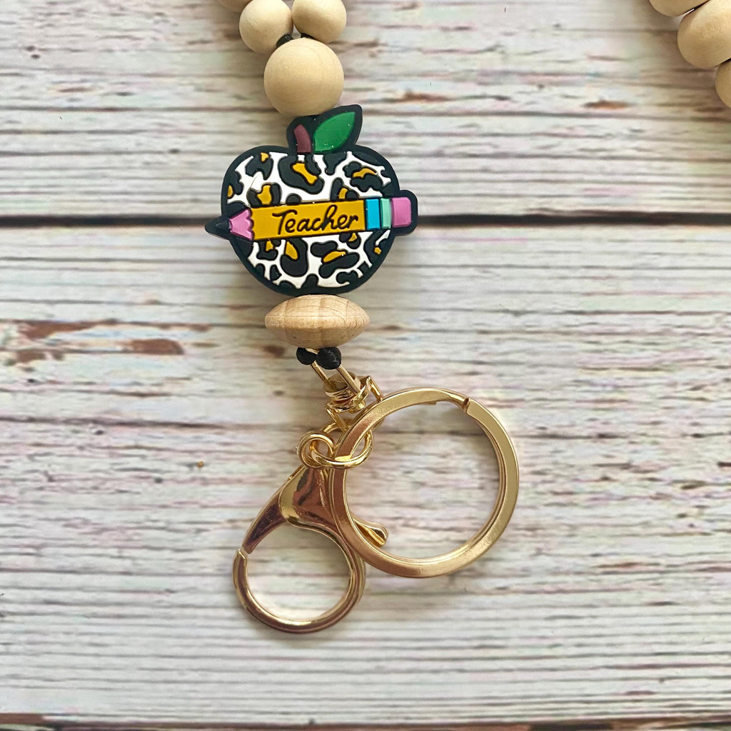 Teacher-themed beaded keychain with a leopard-print apple charm, a yellow 'Teacher' banner, wooden beads, and gold-tone split rings on a whitewashed wood background.