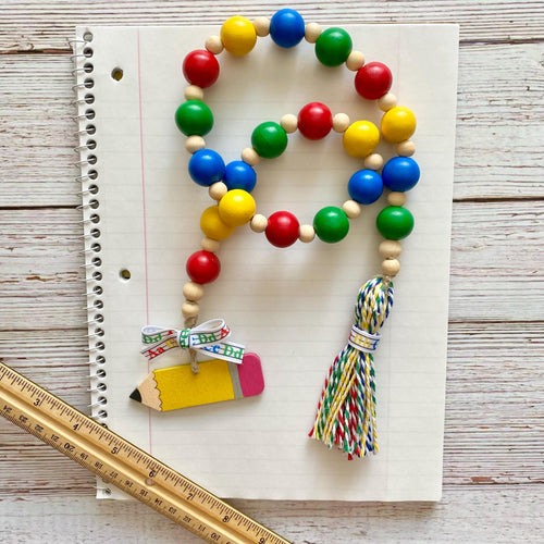A colorful beaded garland with red, blue, green, and yellow round beads, natural wood beads, a yellow pencil charm, and a multicolor tassel, displayed on a lined notebook with a wooden ruler.