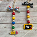 Two personalized school-themed bead garlands hanging on a rustic white-washed wooden background. One garland features a colorful patterned ribbon bow, red, yellow, blue, green, and natural wooden beads, and a yellow pencil charm.