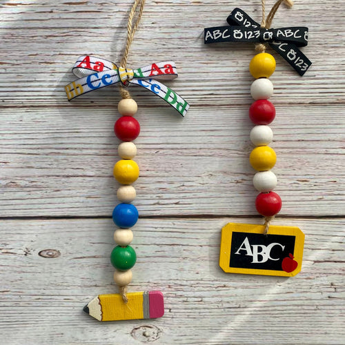 Two personalized school-themed bead garlands hanging on a rustic white-washed wooden background. One garland features a colorful patterned ribbon bow, red, yellow, blue, green, and natural wooden beads, and a yellow pencil charm.